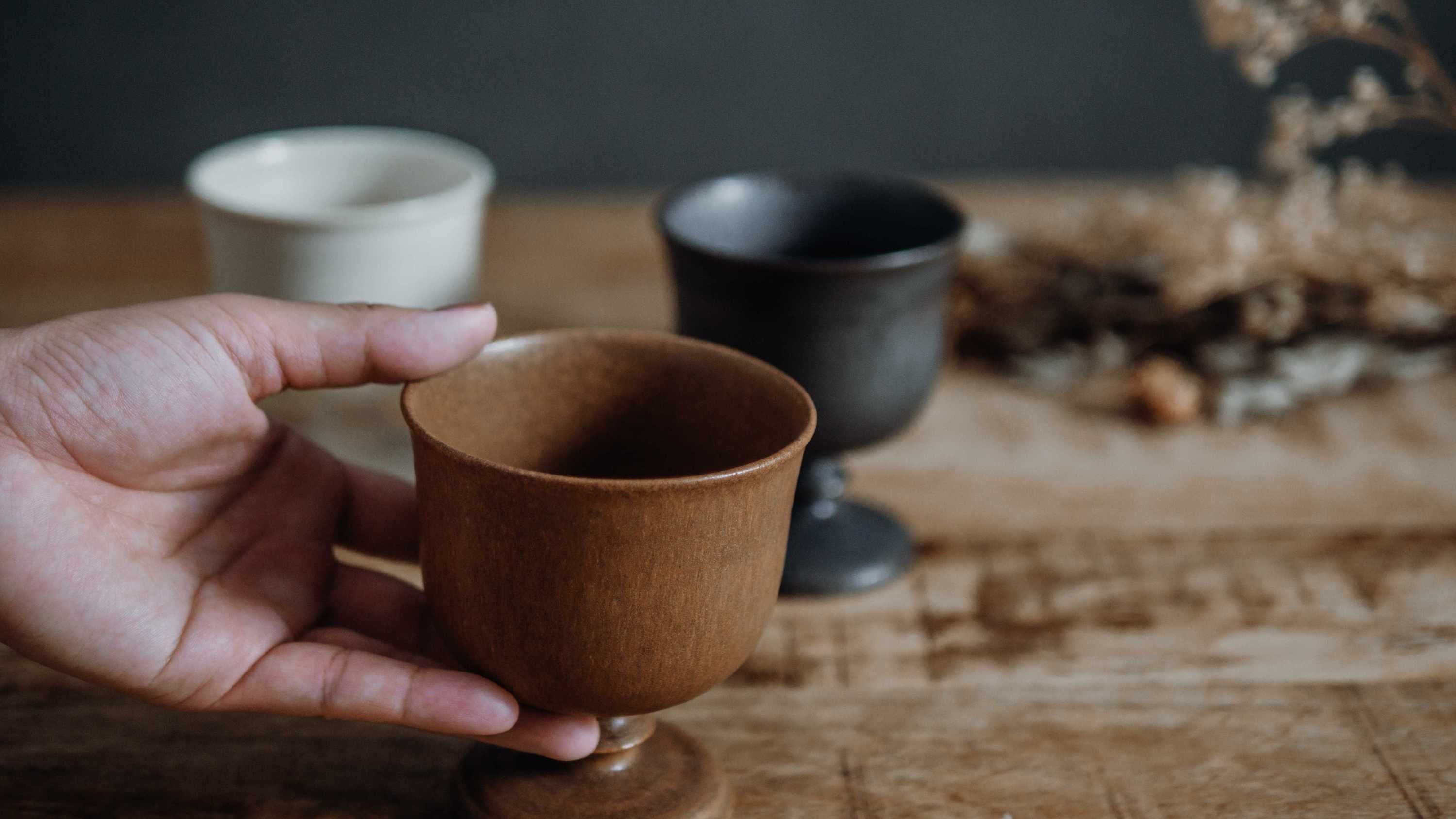 手のひらに乗せた吉田正和さんの茶色の陶器の玉脚カップと背景に並ぶ白と黒のカップたち